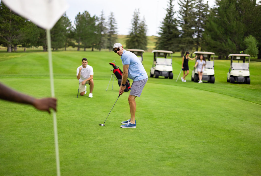 A man squaring up to put a golf ball in the hole with friends in the background talking around their golf carts.