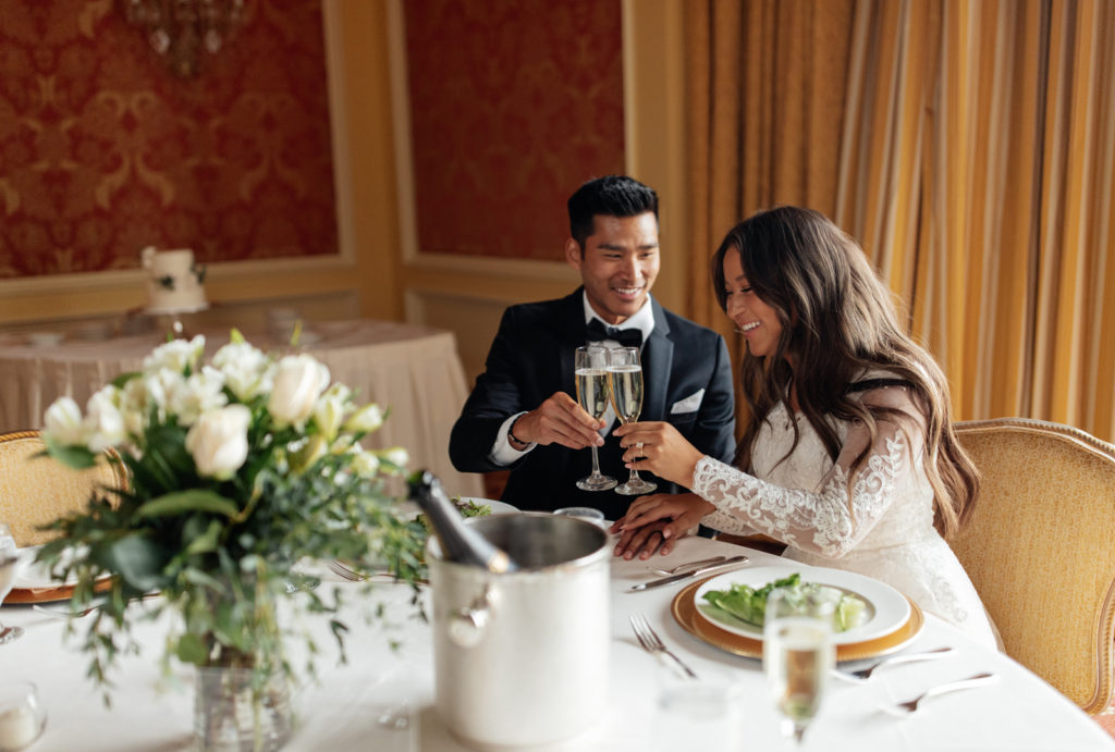 Bride and groom raising a toast with champagne glasses while sitting at a table.