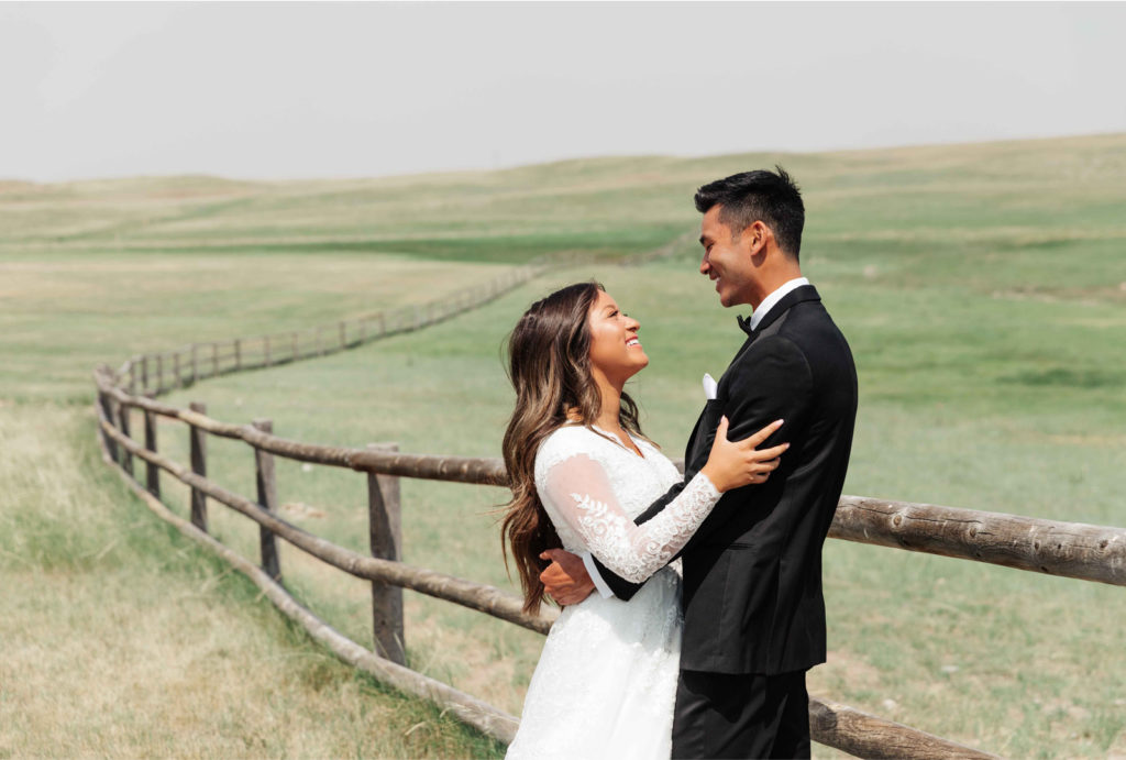 Bride and groom holding each other and smiling at each other while standing in a scenic field.